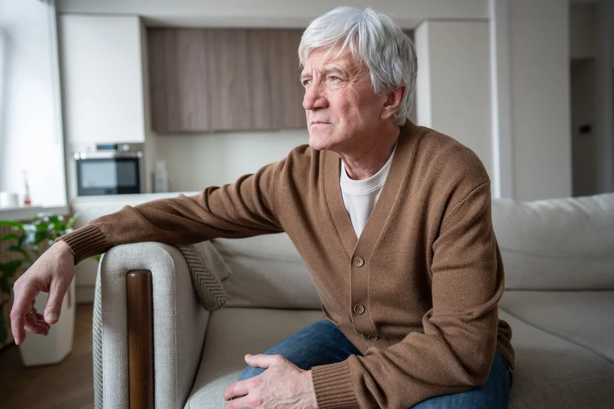 Pensive elderly man looking out the window from a living room.